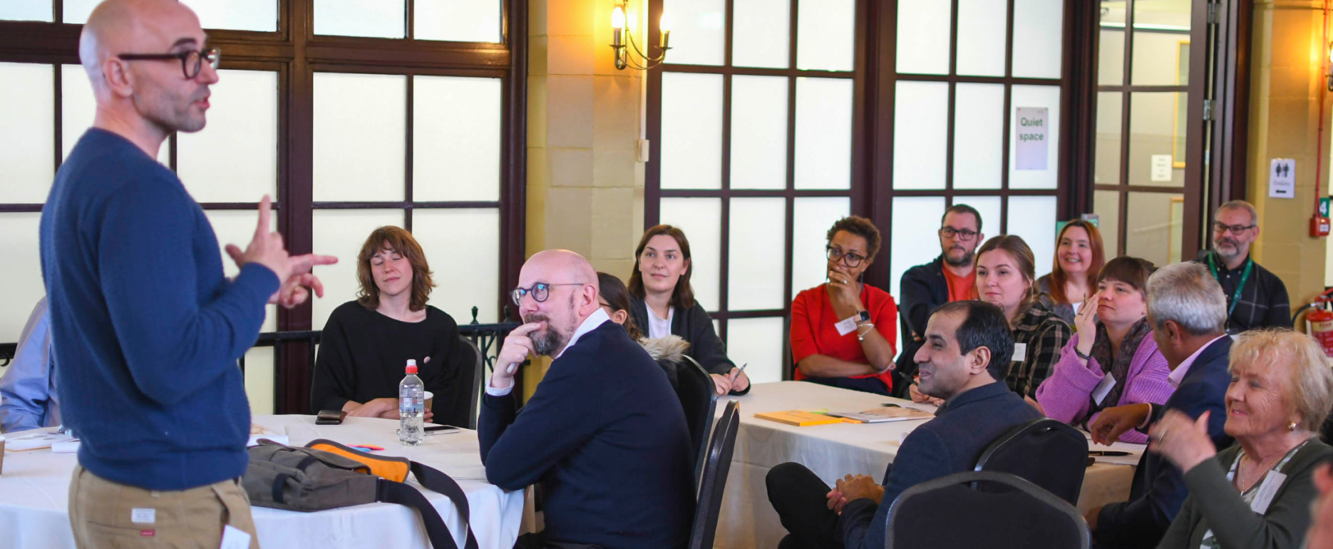 A group of people sit and watch as a man in a blue jumper leads a workshop.