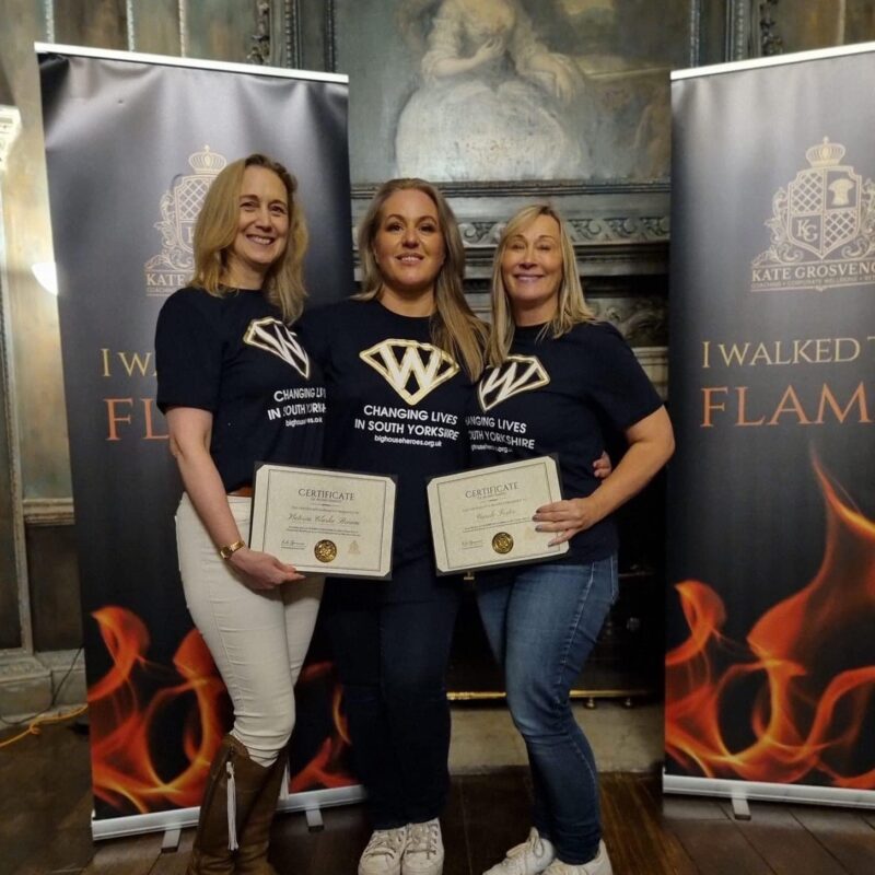 Firewalkers Vicki Clarke Brown (left) and Wentworth Woodhouse’s Fundraising Manager Carole Foster (right) receive their certificates from life coach Kate Grosvenor. The women all wear black t-shirts which read "Changing Lives in South Yorkshire" and smile at the camera.