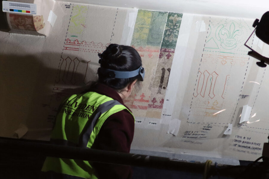 A photo of a person with dark hair in a bun wearing a hi-vis jacket who has their back to the camera. They are looking at papers which show elements of the wall paintings.
