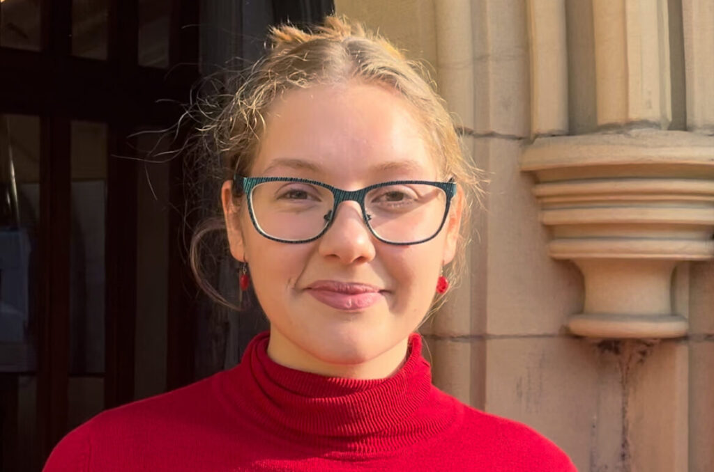 An image of a young woman, wearing a red top, dark rimmed glasses, with a pale stone column in the background