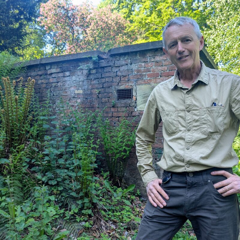 Simon Gill, Head of Heritage (Buildings and Landscapes) stands outside Wentworth Woodhouse's dilapidated Ice House. It is a small brick building surrounded by vegetation.