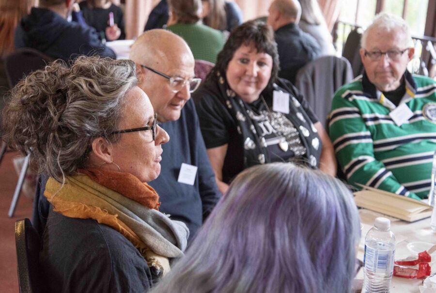 A group of people sat around a table look on in interest as one of them speaks to the group.