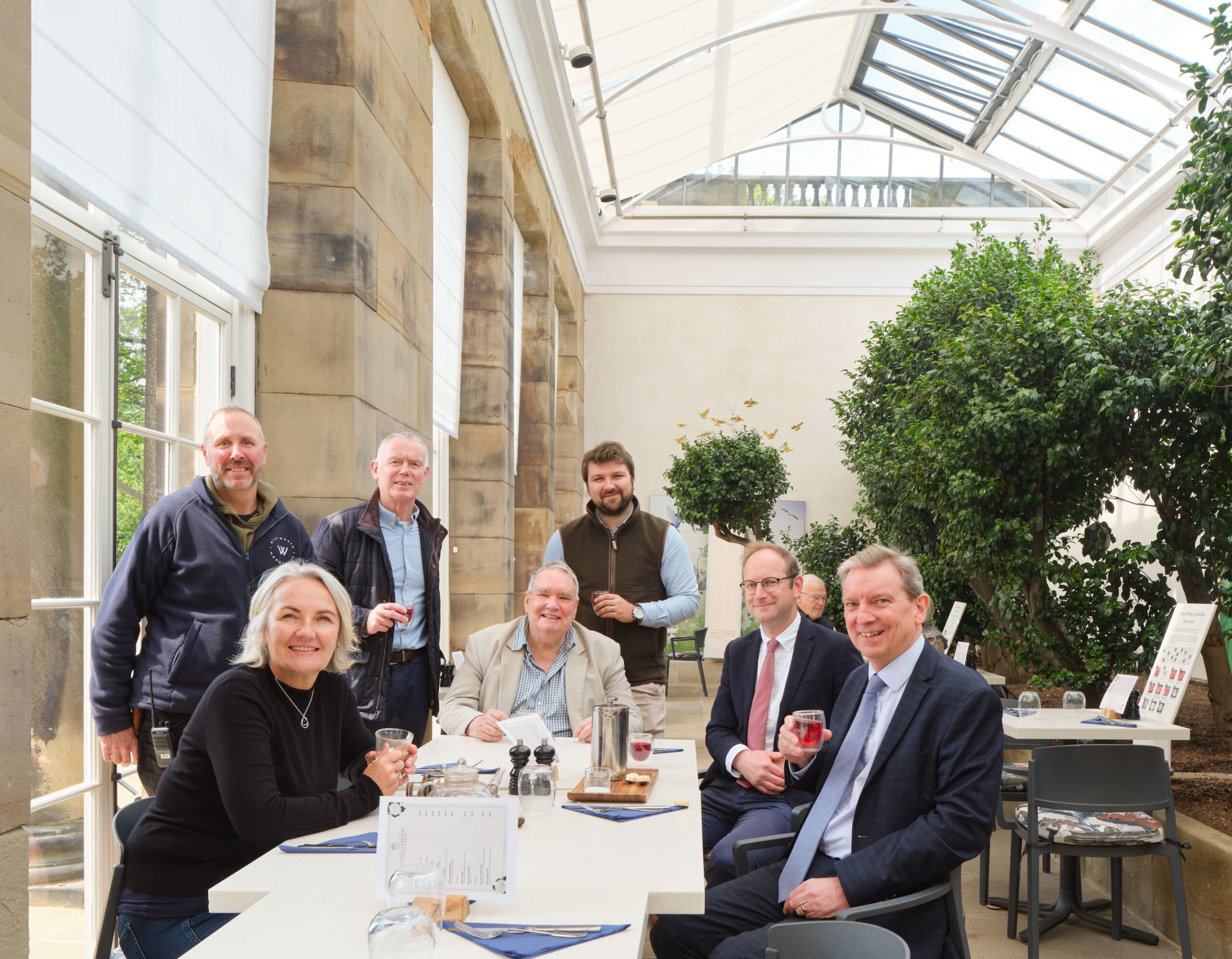 Seven people sitting around a table in a naturally brightly lit terrace. The terrace is part of the renovated Camellia House.