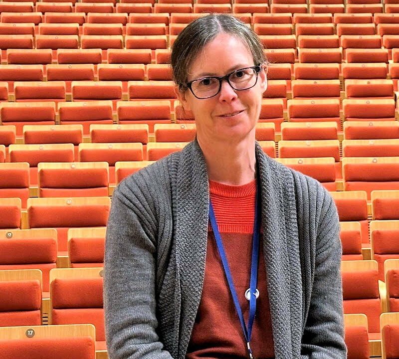 Woman sat in front of orange theatre seats. She wears a grey cardigan and red top and is facing the camera