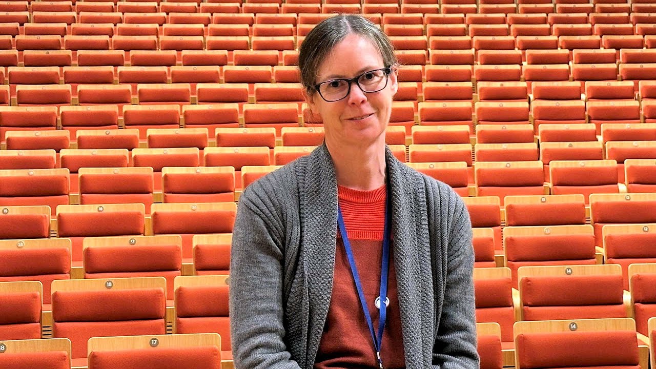 Woman sat in front of orange theatre seats. She wears a grey cardigan and red top and is facing the camera