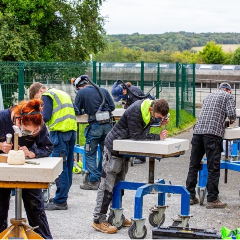 Eight apprentices, or recently qualified stone masons, are showcasing their creative talents. They have tablets of stone on tables outdoors and are carving them.