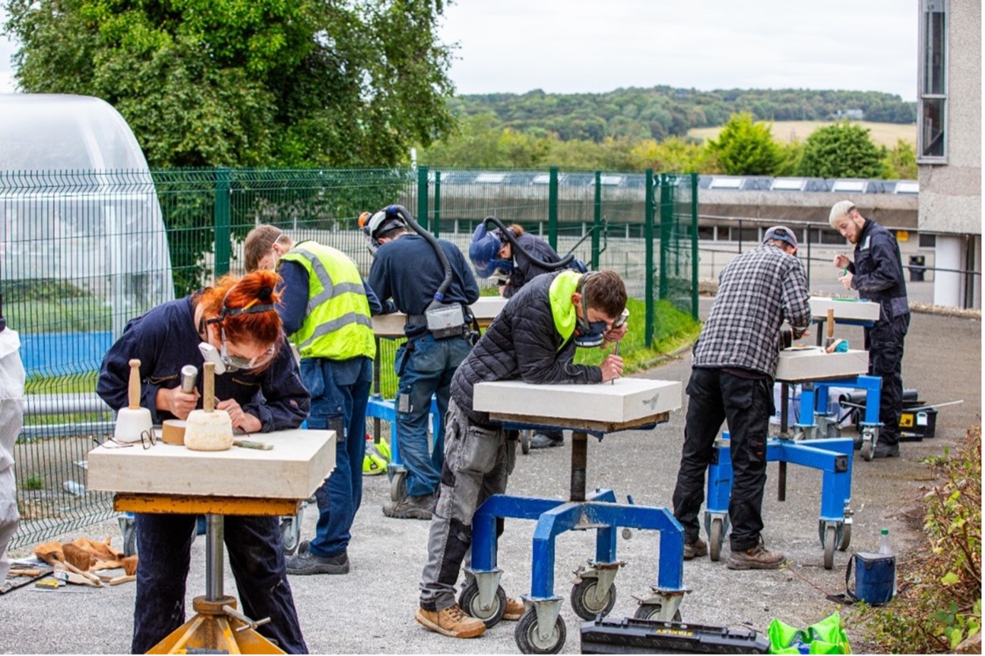Eight apprentices, or recently qualified stone masons, are showcasing their creative talents. They have tablets of stone on tables outdoors and are carving them.