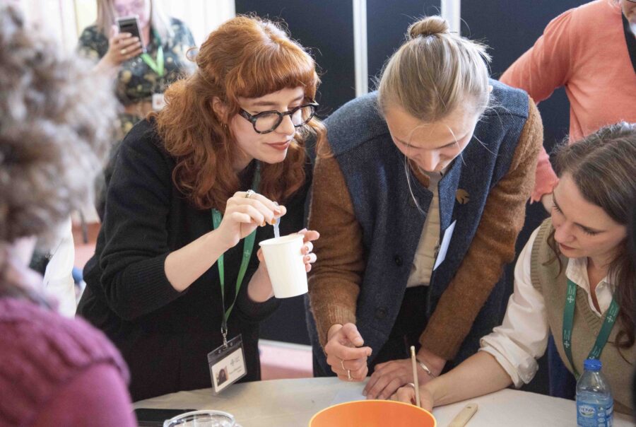 A group of people gather around a table at an event, they are working together on something, one person writing, whilst another gathers materials.