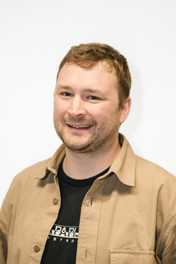 Man facing the camera and smiling against a white background. He is wearing a beige coloured shirt and black vest top