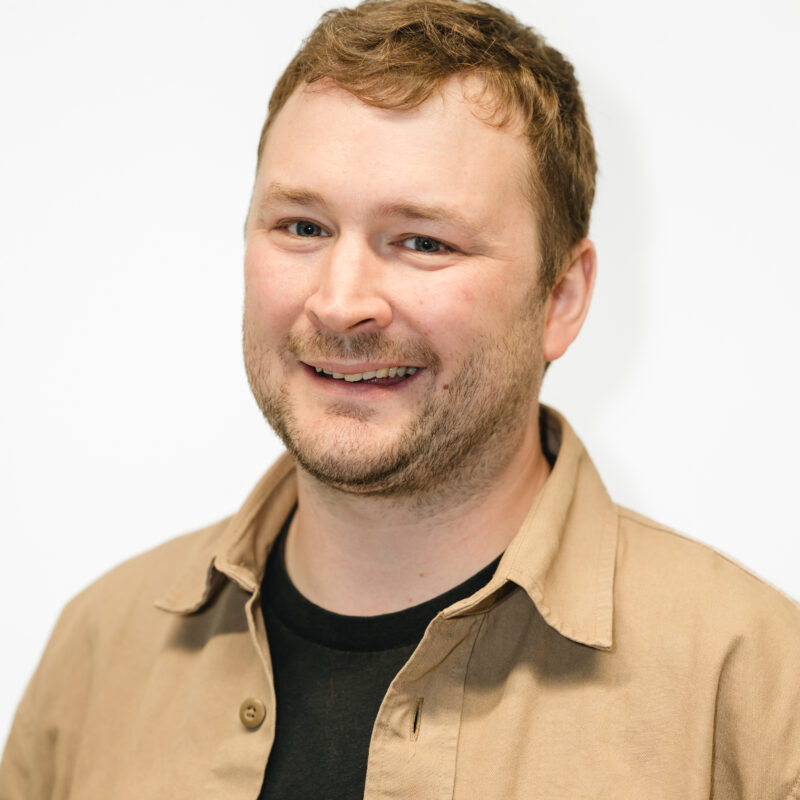 Man facing the camera and smiling against a white background. He is wearing a beige coloured shirt and black vest top