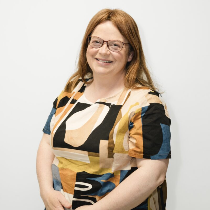 Martina smiling at the camera, wearing a patterned dress and standing against a white background