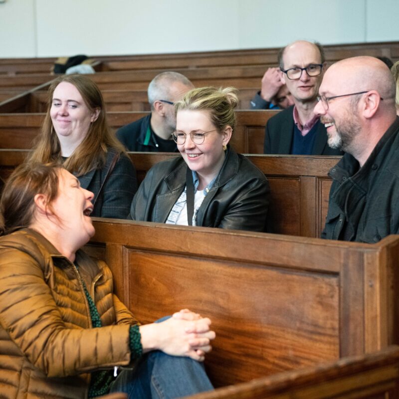 A group of people sit on church pews, they are talking together, one person laughs heartily as the others look on smiling.