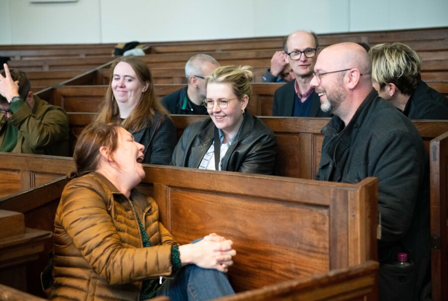 A group of people sit on church pews, they are talking together, one person laughs heartily as the others look on smiling.