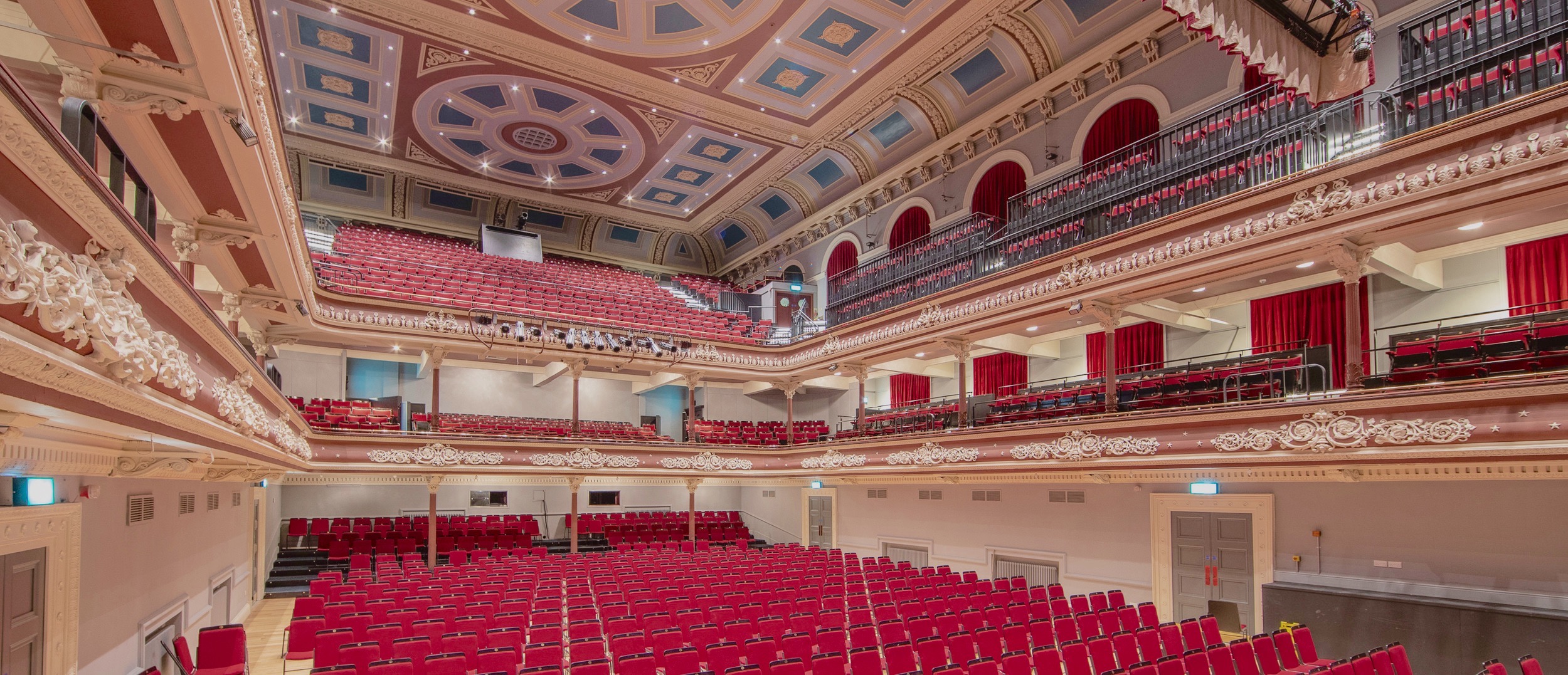 Wide photograph of the interior of St George's Hall, with ornate moulding on the ceiling and balconies, red velvet seats fill the floor and balconies.