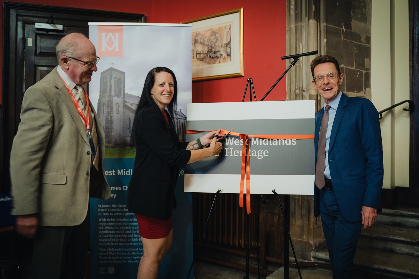 Three people stand around a sign that reads 'West Midlands Heritage Trust'. The sign has an orange ribbon tied around it, and one person is cutting the ribbon as part of an opening ceremony.
