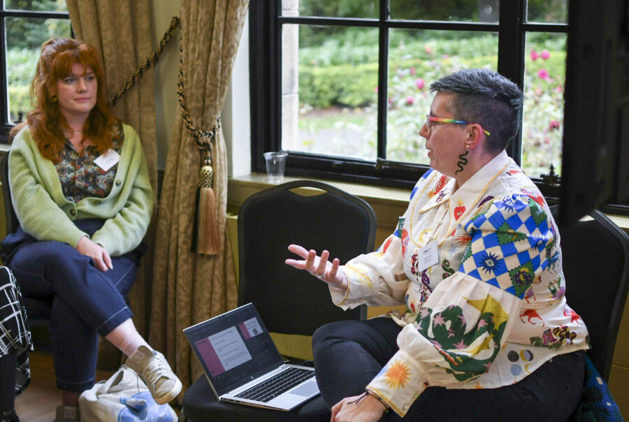 A non-binary person in a colourful patterned shirt leads a conference talk, audience members look on engaged.