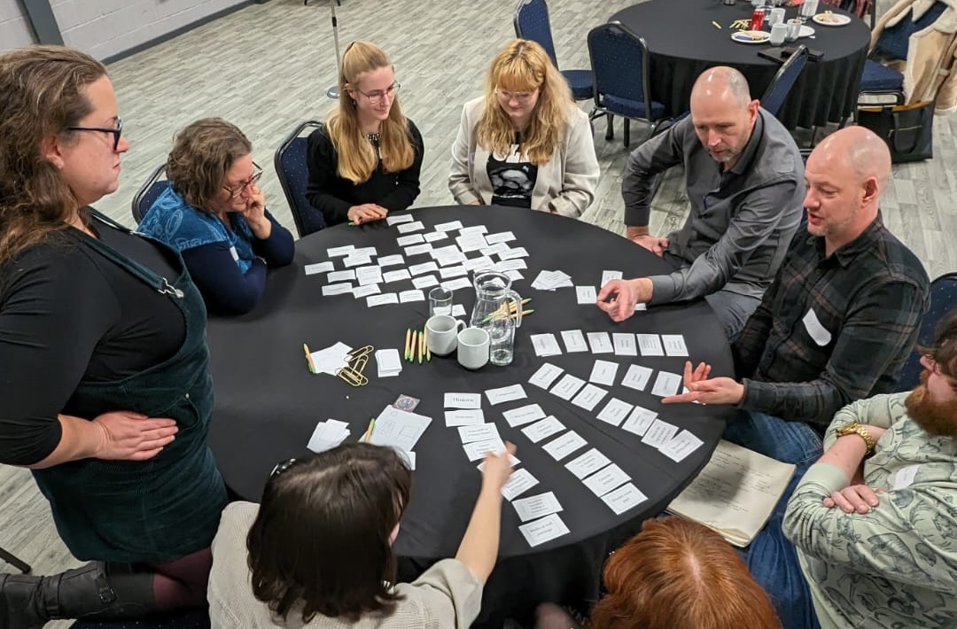 A group of people stand and sit around a round table. They are working together to arrange a series of rectangular cards in order.