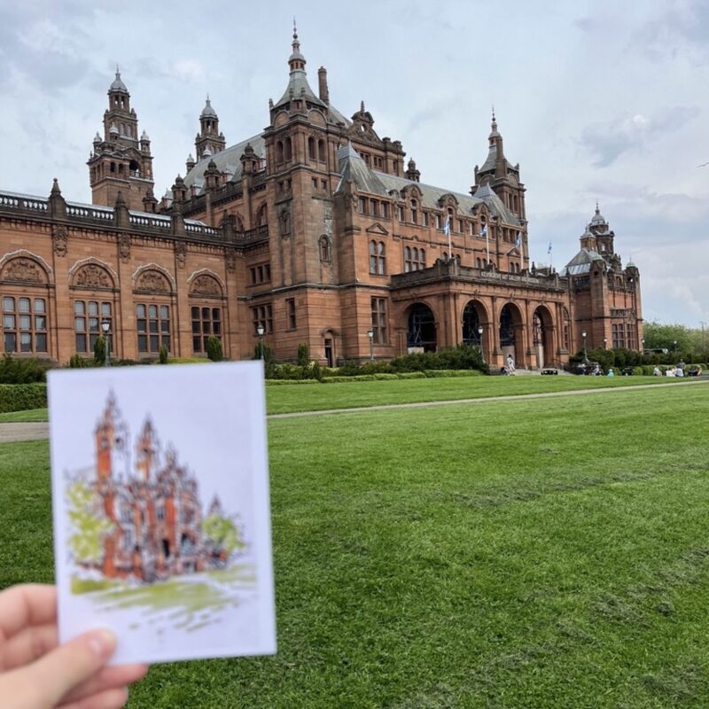 Photograph of Kelvingrove Museum in Glasgow. In the foreground, out of focus, is a hand holding a small illustration of the museum.