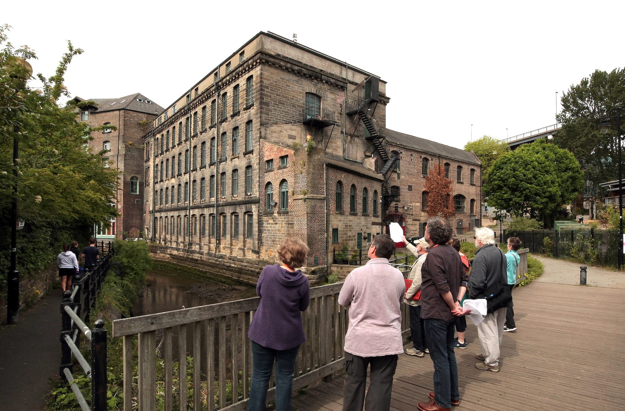 A group of people stand on a wooden bridge, looking up at a brick building with many windows.