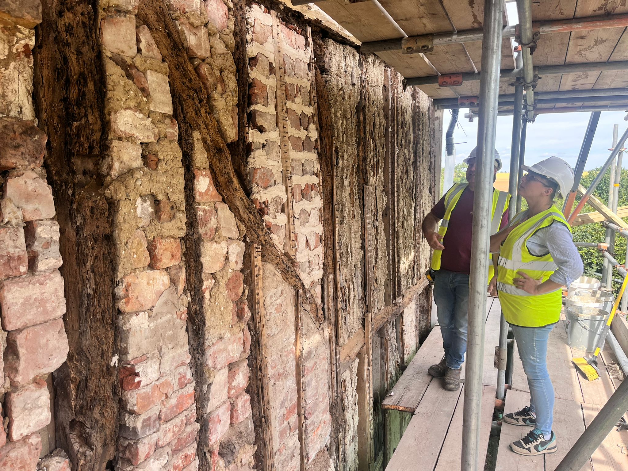 Two people in hi-viz jackets and hard hats stand on scaffolding inspecting the exposed bricks and timber of the chapel.