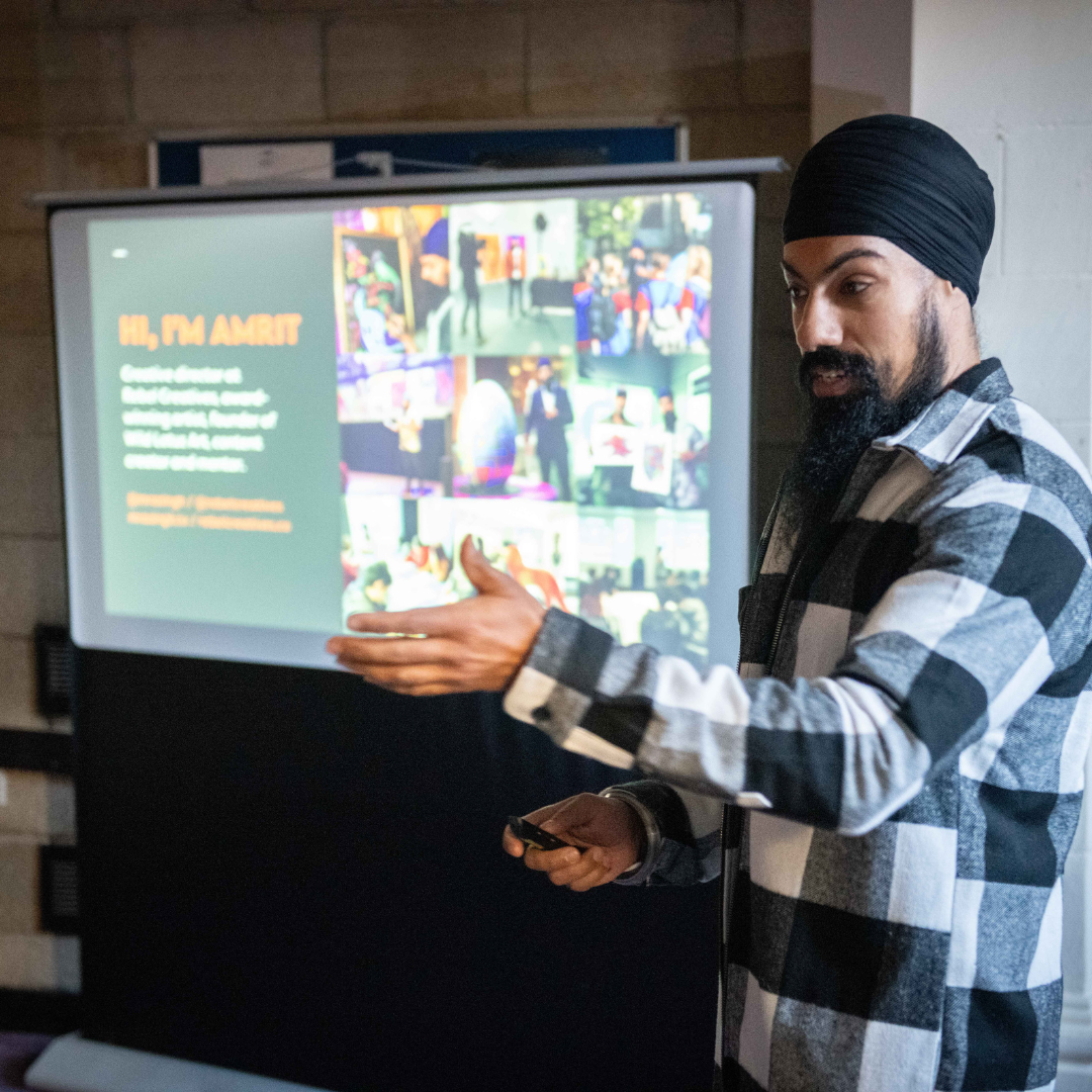 Photograph of a man in a check shirt, with a turban headwrap giving a presentation, he is gesturing to a screen behind him.