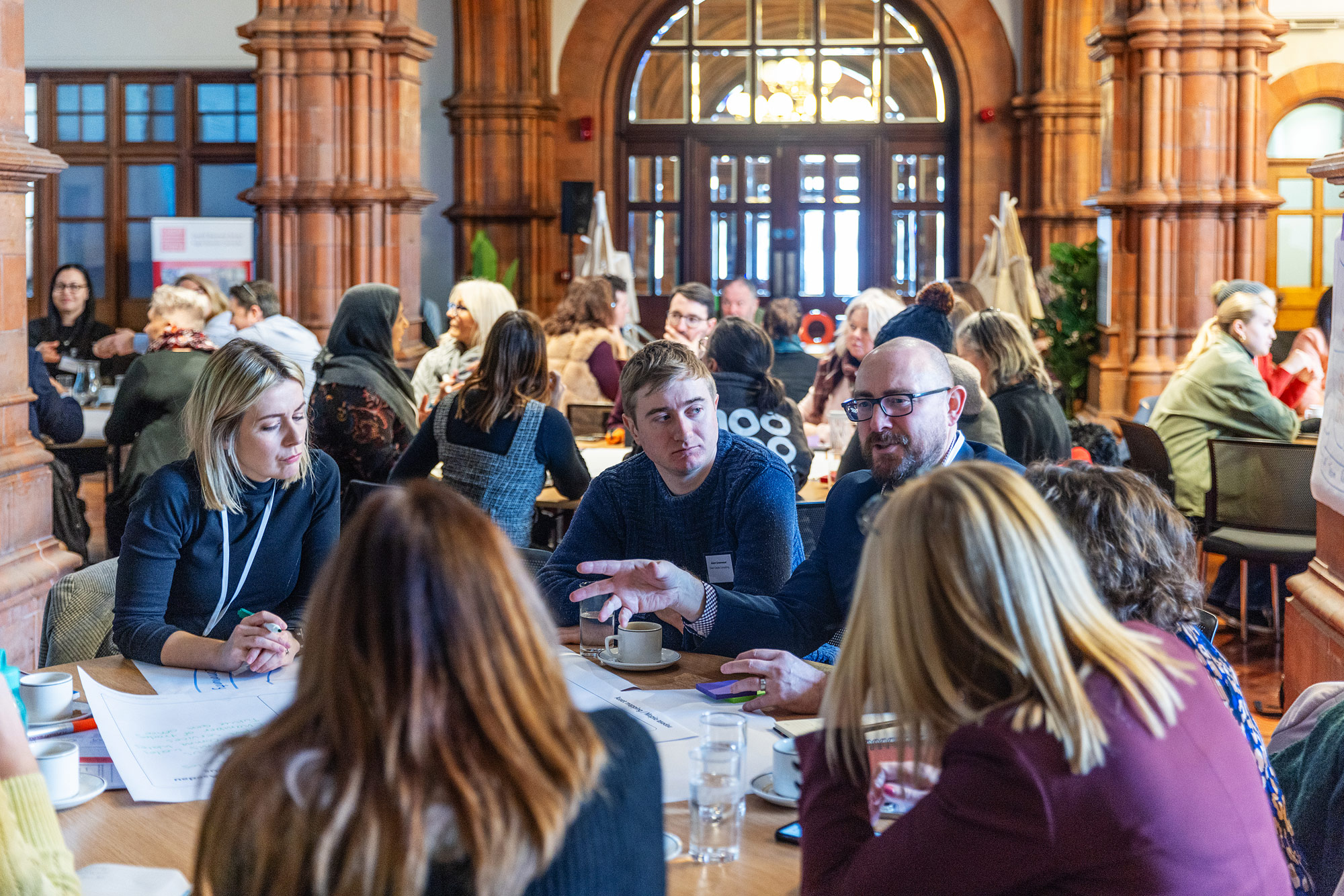 A photo from Cwmpas' 'Hackathon', a group of people sit around a table in a busy hall, one is speaking and the other people are listening appearing very engaged