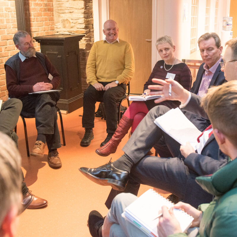 A group of people sit together on chairs arranged in a circle, they are having a discussion and all look engaged in what the speaker is saying.