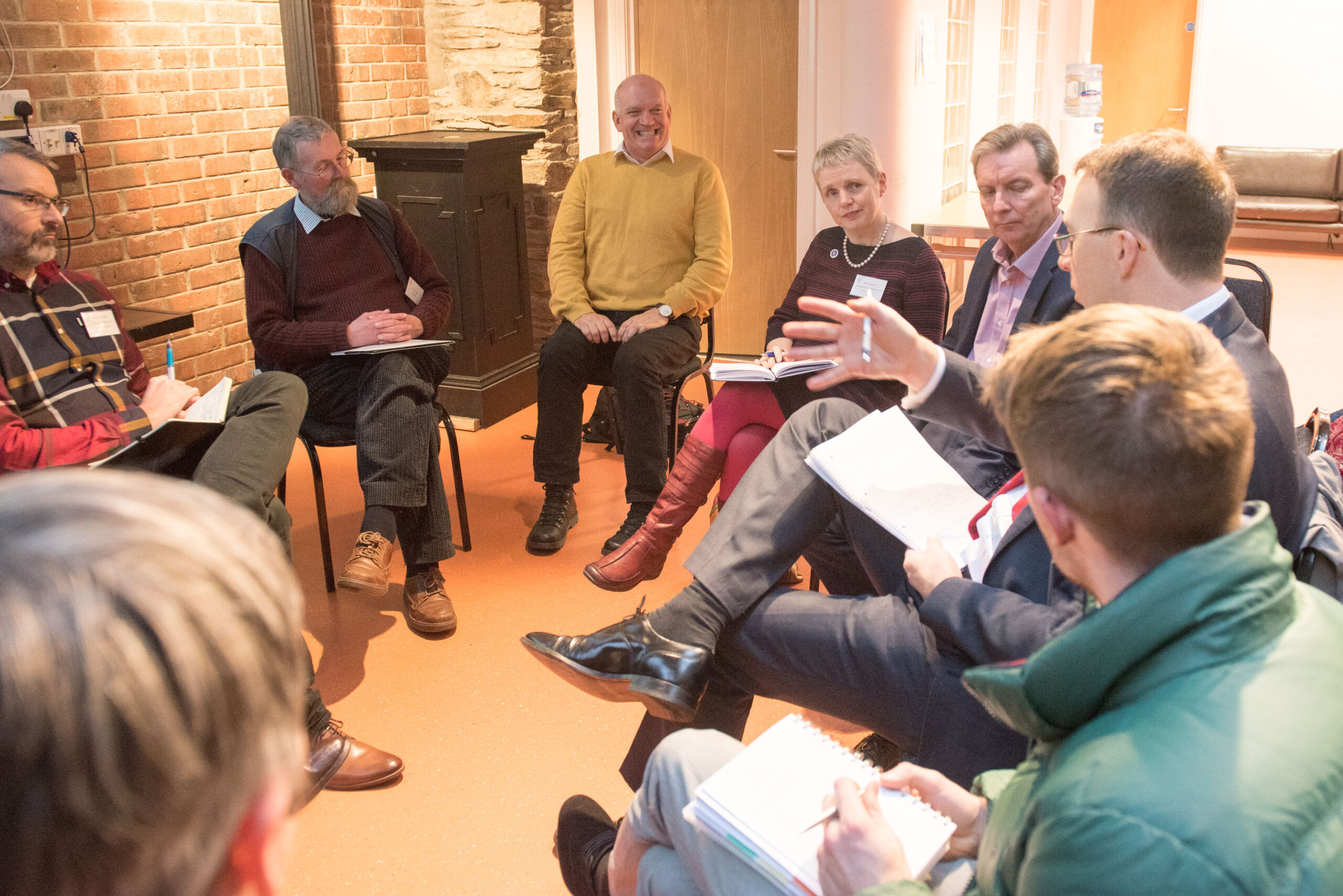 A group of people sit together on chairs arranged in a circle, they are having a discussion and all look engaged in what the speaker is saying.