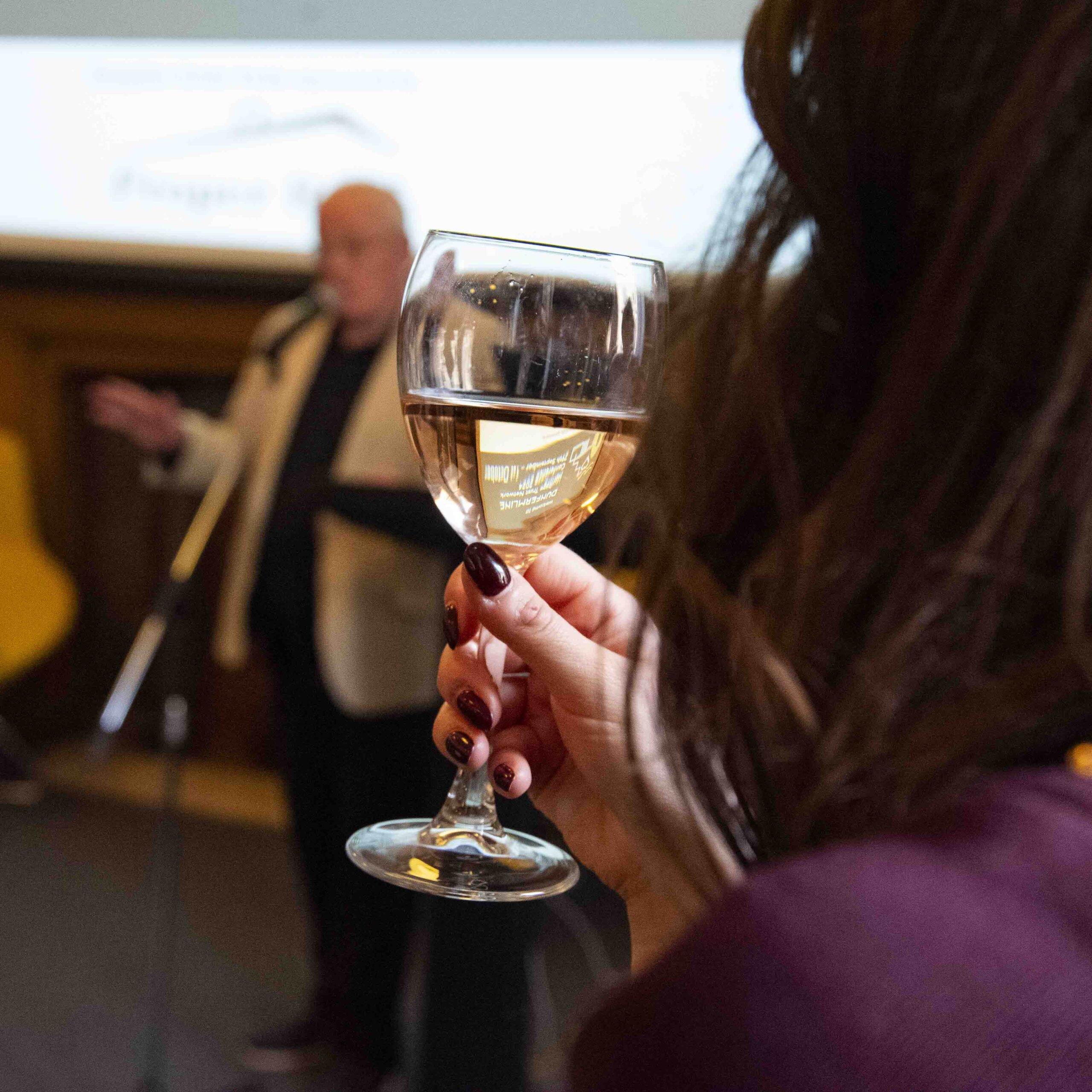 Photograph of a person holding a drink in a wine glass, out of focus in the background a talk is happening.