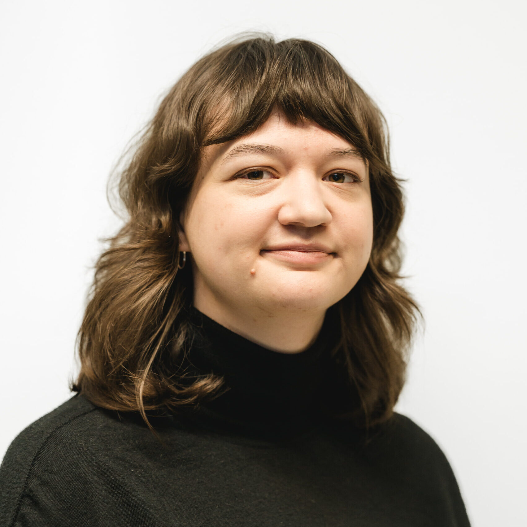 Samya wears a black turtleneck top and smiles at the camera, she has medium length brown hair and is shown from the shoulders up against a white background.
