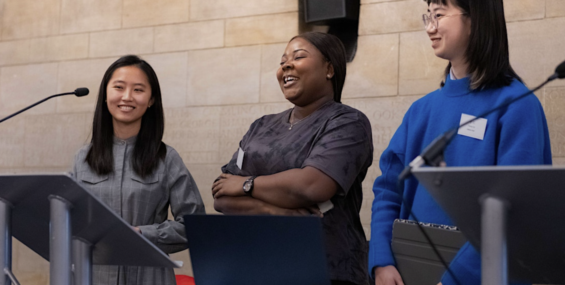 three young people stand on a stage behind a podium, they are smiling and giving a speech