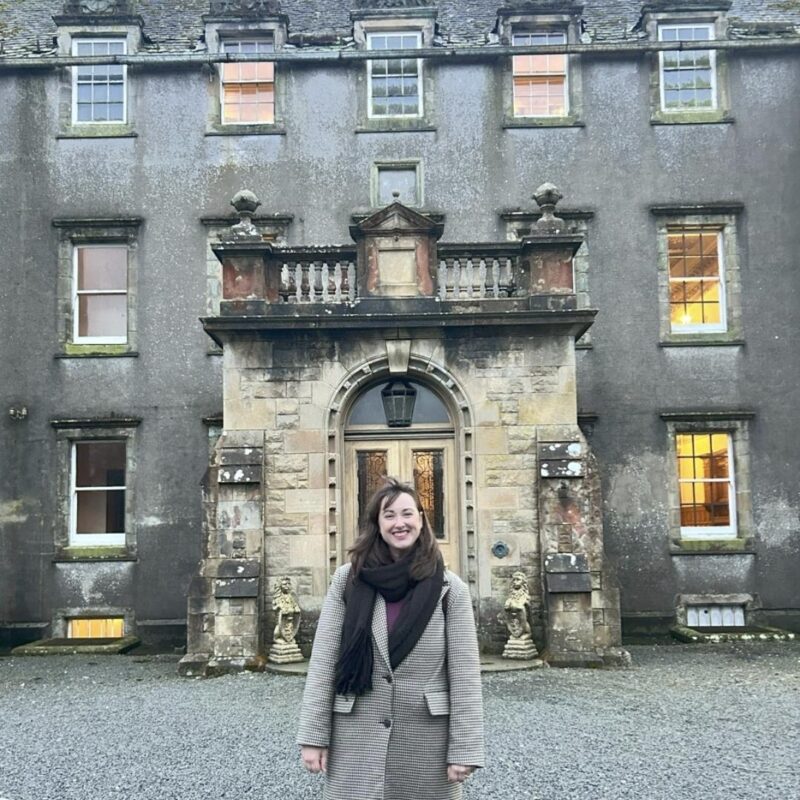 A dark haired lady in a full length coat stands in front of the house. There is a large entrance stone porch and the building is three storey with many windows.