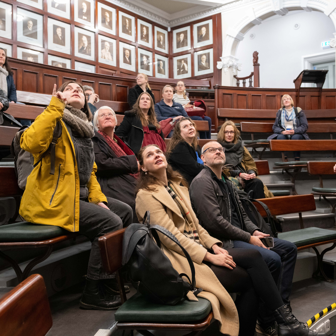 Photograph of a tour group sat in an historic building, they are looking at features and pointing them out to each other.