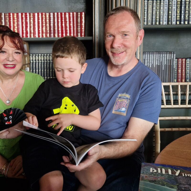 A man and woman sit in front of shelves of books. On their lap sits a young boy who is reading a large paperback book.
