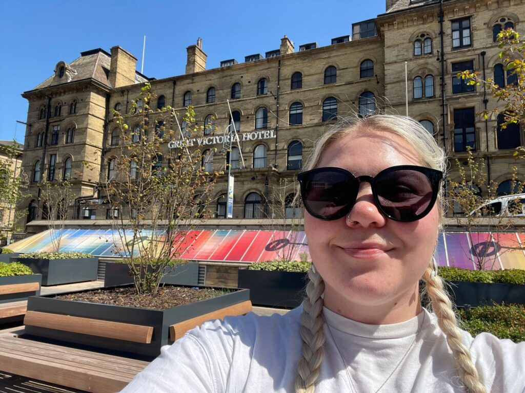 Izabella outside the Great Victoria Hotel in Bradford on a sunny day. She is wearing black sunglasses a white top and has blonde hair