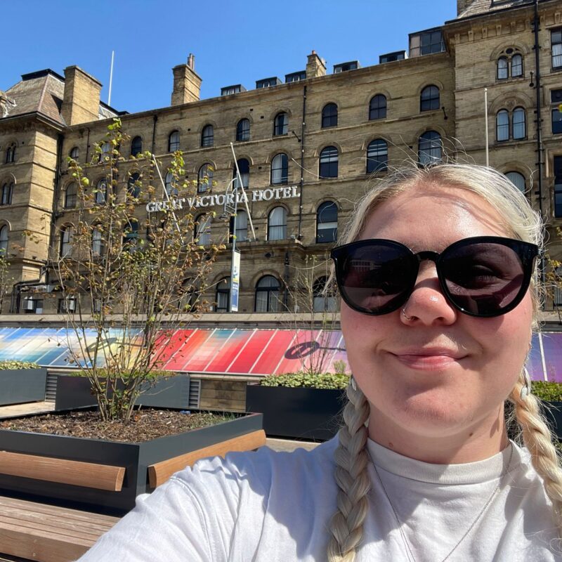 Izabella outside the Great Victoria Hotel in Bradford on a sunny day. She is wearing black sunglasses a white top and has blonde hair