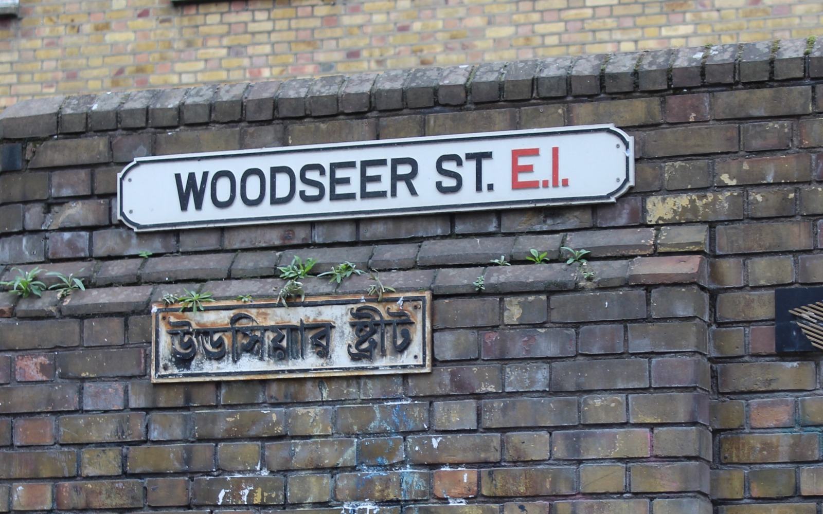 Brick wall with a sign reading 'Woodseer St. E.I.' Underneath there is a sign in another language.