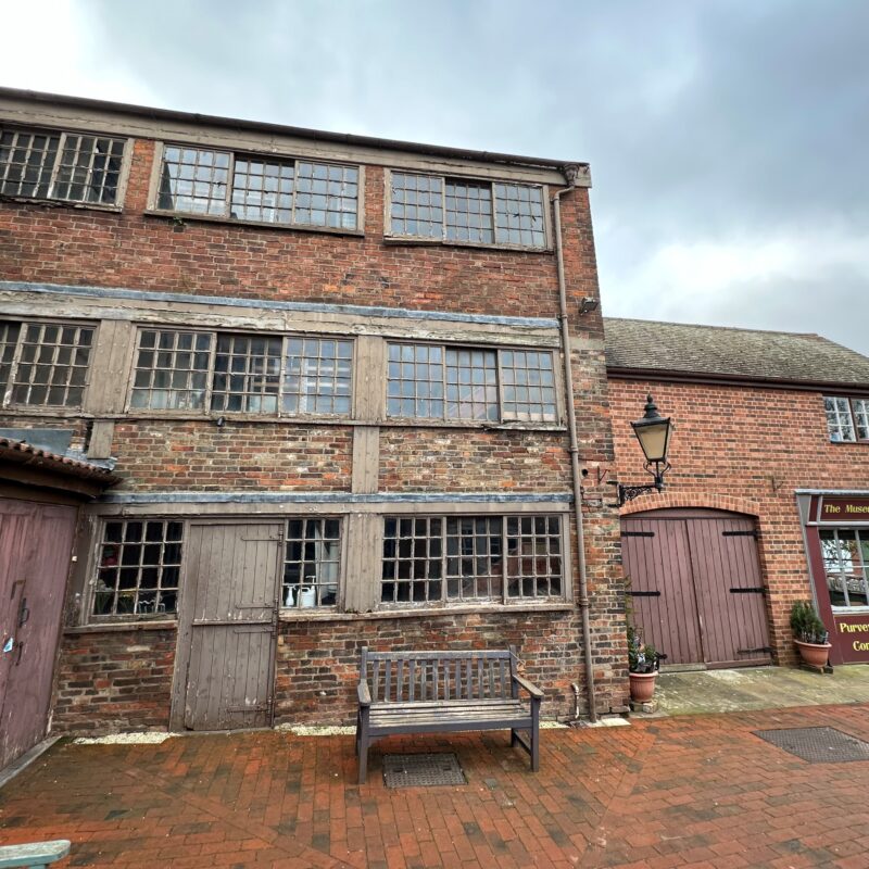 the pin factory, a red bricked three story building, it sits in a paved redbrick court yard and a bench site outside the building.