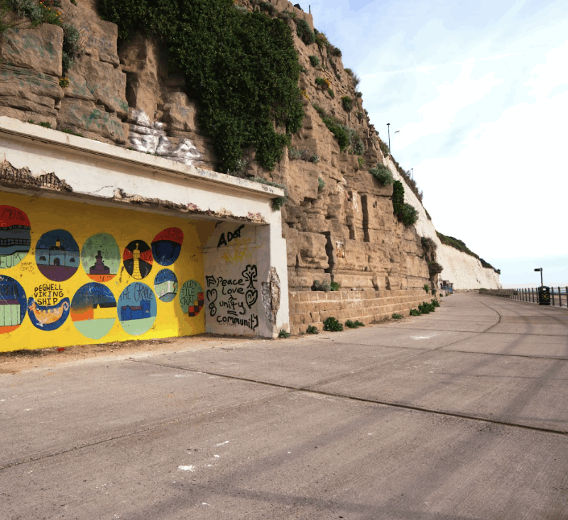 A photograph of one of the East Cliff Shelters. It is a recessed space in the cliff. It's back wall is painted bright yellow with colourful decorations.