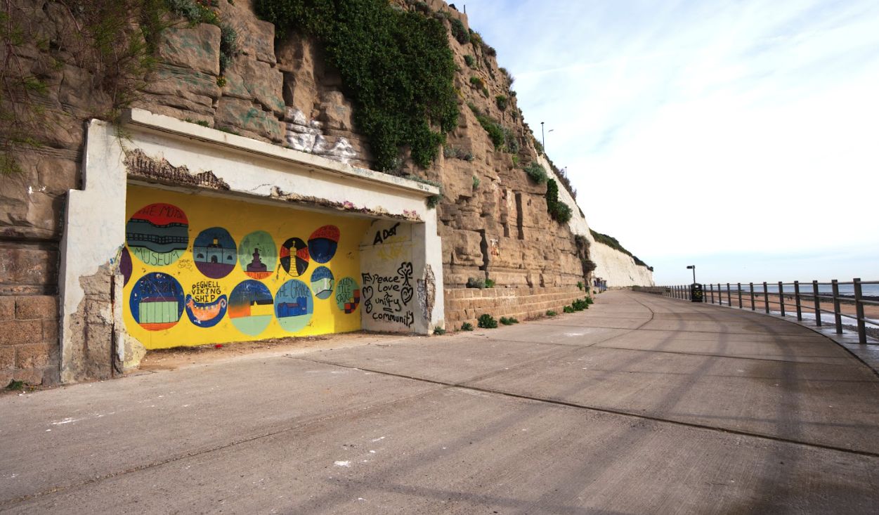 A photograph of one of the East Cliff Shelters. It is a recessed space in the cliff. It's back wall is painted bright yellow with colourful decorations.