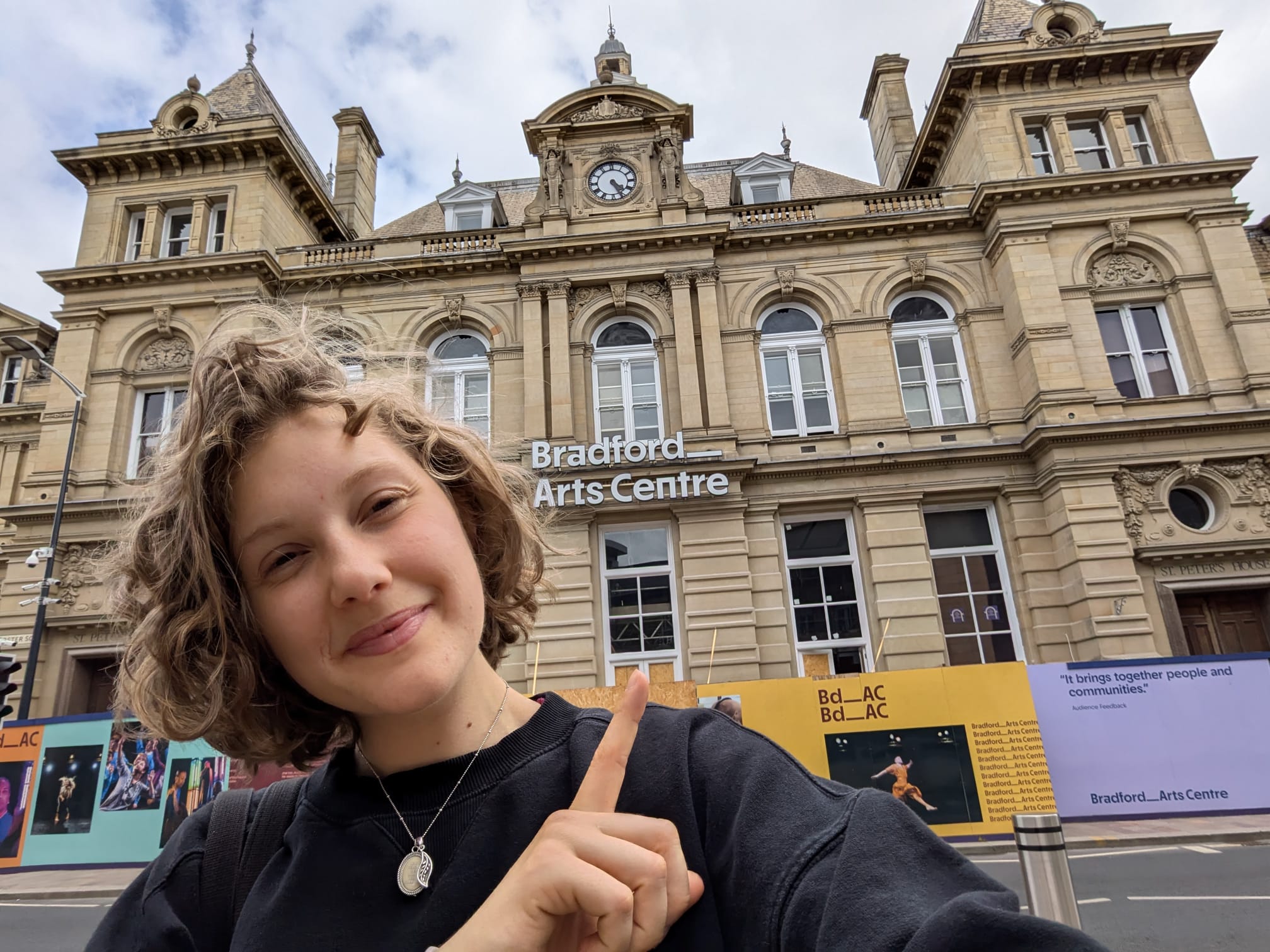 Sophie, a young white woman, is smiling and pointing towards the Bradford Art Centre building behind her.