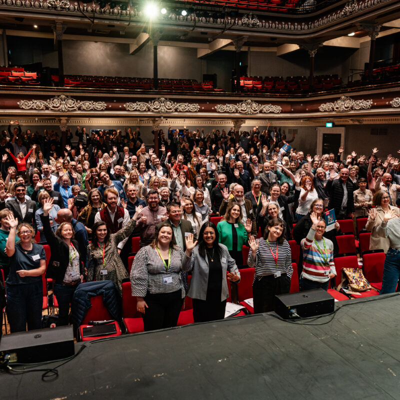 a large group of people in a theatre style setting waving to the camera.