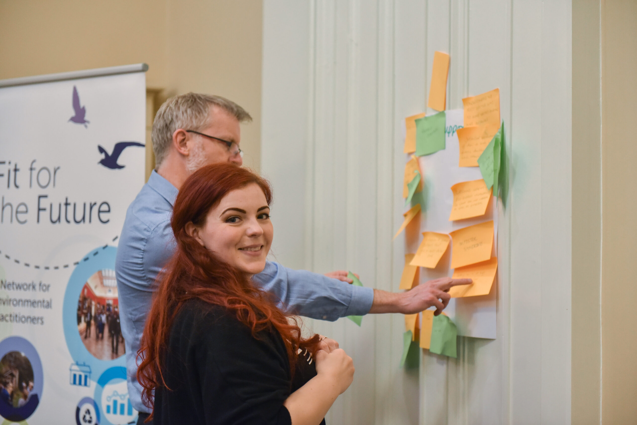 A young woman and a grey haired man are planning something using sticky notes on a wall. The young woman is looking towards the camera smiling.
