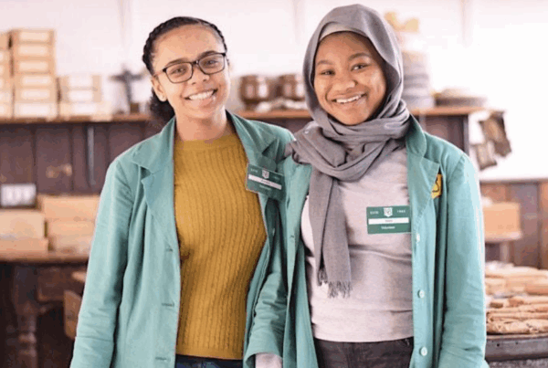 Two young people are smiling at the camera, both are wearing pale green jackets, one is wearing glasses, one is wearing a grey headscarf. The background shows brown paper packages.