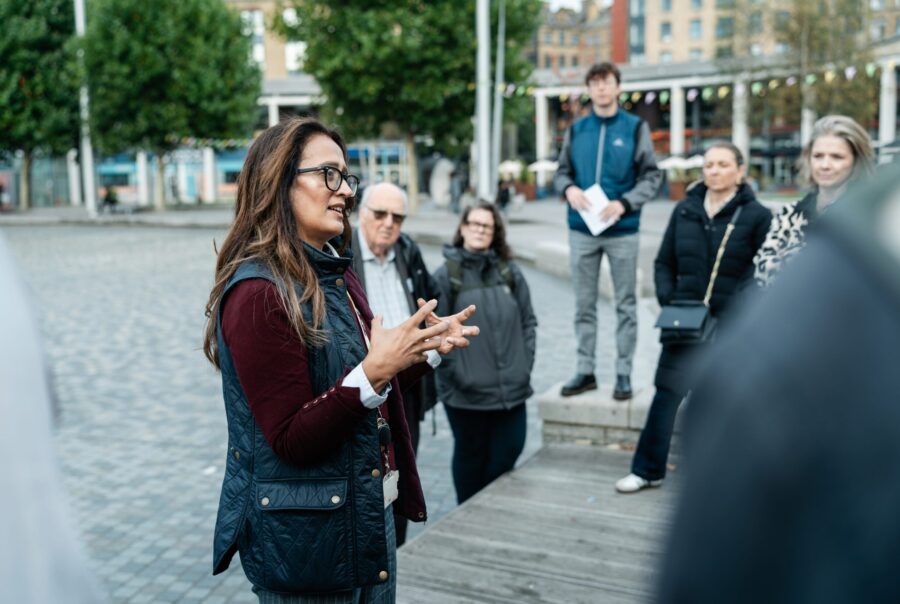 A group of people are looking at one person in the centre of the image who is leading a walking tour. The background is grey street pavement.