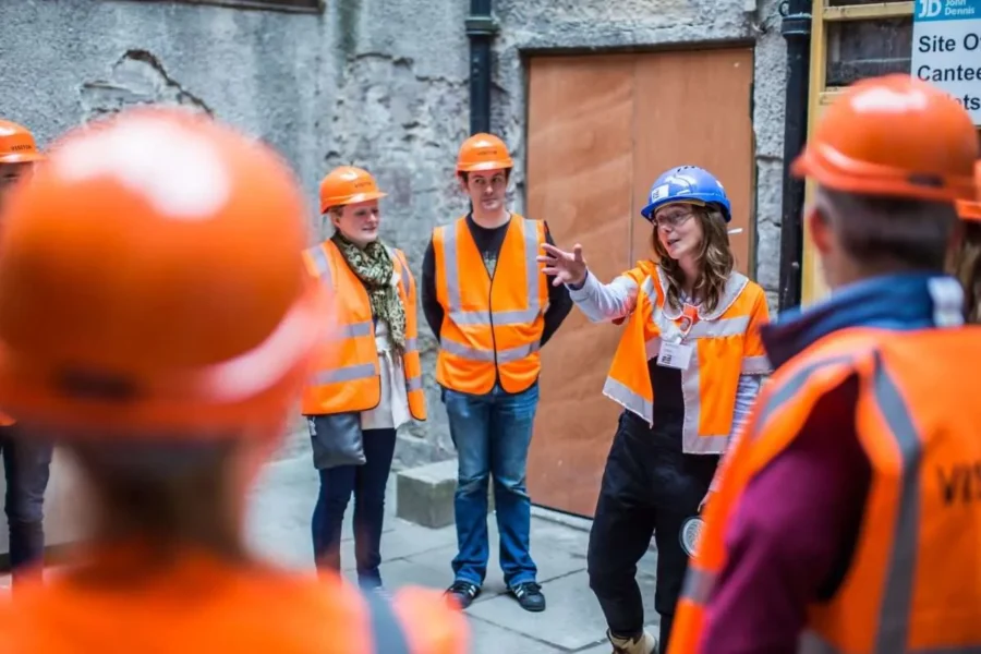 A group of people in orange high-vis vests and hard hats listen to a woman carrying a torch.