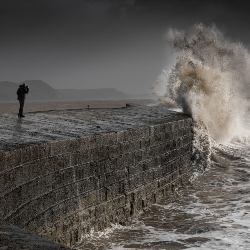 Large white wave crashing against a stone barrier on a headland, with a person in the mid ground taking a photograph of the wave