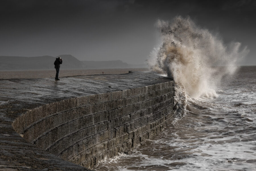 Large white wave crashing against a stone barrier on a headland, with a person in the mid ground taking a photograph of the wave
