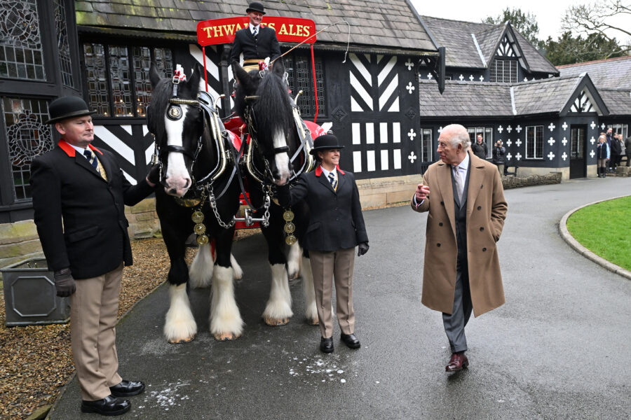 King Charles III stands outside Samlesbury Hall. Next to him is a cart and two shire horses and three people in black jackets and bowler hats supporting the horses.
