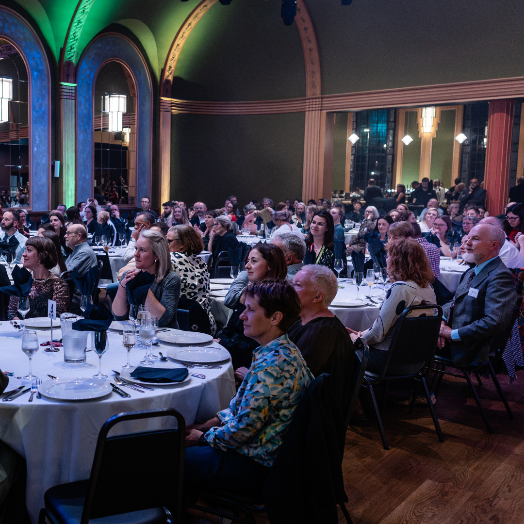 Photograph of a conference dinner in a historic ballroom, delegates are looking at a speaker out of shot.